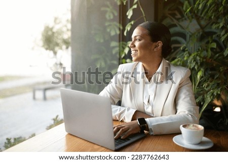 Glad millennial african american businesswoman in white suit using laptop, looking at window in cafe with green plants interior. Coffee break, startup, ad and offer, business, work remotely