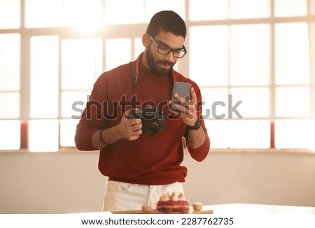 Photographer handsome hispanic young man taking photo of tasty colorful beautiful dessert, using digital camera, holding smartphone, checking trends on social media, sunlight behind his back Royalty-Free Stock Photo #2287762735