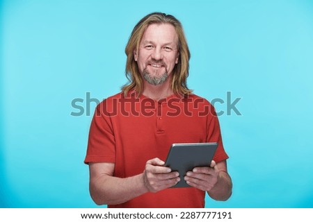 Portrait of mature man in red shirt smiling at camera on blue background and using digital tablet