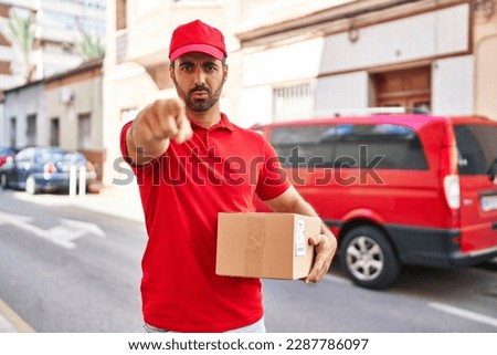 Young hispanic man with beard wearing delivery uniform and cap holding box pointing with finger to the camera and to you, confident gesture looking serious 