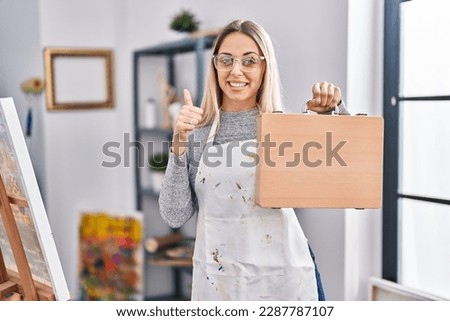 Young blonde painter woman holding wooden case at art studio smiling happy and positive, thumb up doing excellent and approval sign 