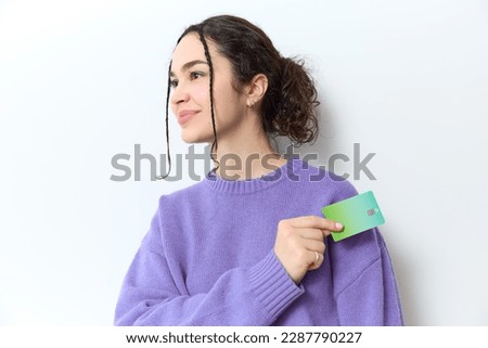Gen z woman with curly braids and ponytail in purple sweater smiles happily while holding a green credit card to pay in her hand on white backdrop in studio. Shopping online, banking, nfc concept.