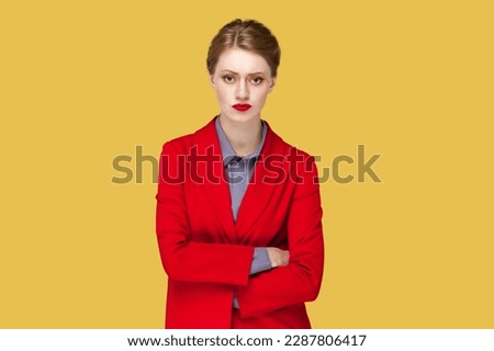 Portrait of serious strict woman with red lips standing standing with crossed arms, looking at camera, having bossy expression, wearing red jacket. Indoor studio shot isolated on yellow background. Royalty-Free Stock Photo #2287806417