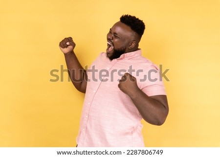 Portrait of positive satisfied man wearing pink shirt raised clenched fists, being happy of winning, expressing triumph, screaming happily. Indoor studio shot isolated on yellow background. Royalty-Free Stock Photo #2287806749
