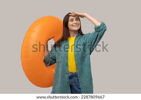 Satisfied woman looking far with hand near eyes and holding orange rubber ring, waiting to travel to tropical country, wearing casual style jacket. Indoor studio shot isolated on gray background. Royalty-Free Stock Photo #2287809667