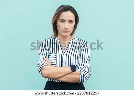 Portrait of serious middle aged woman wearing striped shirt standing with crossed arms and looking at camera with strict bossy expression. Indoor studio shot isolated on light blue background. Royalty-Free Stock Photo #2287812257