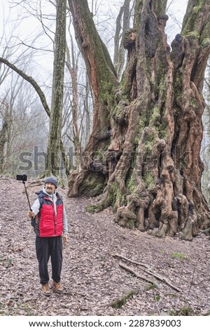 Senior asian man taking selfie on phone with old poplar tree in relic forest. Full length. Front view.Paradise Place Arboretum Park, Sochi, Russia.Populus alba. Technologies, elderly life concept