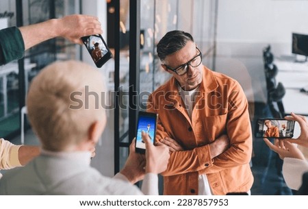 Caucasian businessman in classic eyewear for vision protection posing during leadership meeting in office interior, unrecognizable employees making photos of confident mentor using cellular gadgets