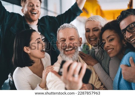 Cheerful male employee making positive selfie photos during day brainstorming with successful colleagues, happy group of professional smiling at camera while shooting video vlog about business