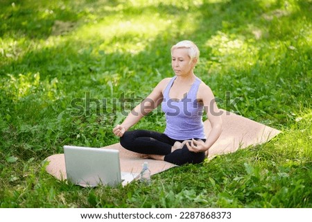 Happy athletic middle aged woman doing online yoga outdoors in a city park in a lotus position sitting on a mat, a bottle of water is standing nearby. The concept of online training, stretching, yoga.