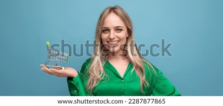 horizontal photo of stylish european woman shopper in green shirt with supermarket trolley on blue studio background with free space