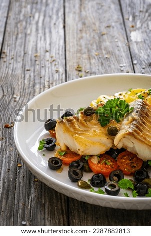 Cod in Italian style on tomatoes and black olives with baked potatoes on wooden table