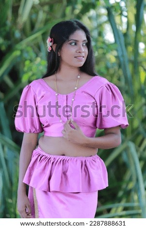 A beautiful young woman in a pink dress. Facing right, in a green environment, with folded arms.