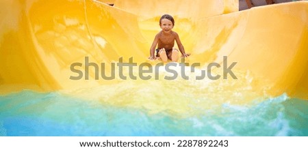 Cheerful blue-eyed kid on a water slide in the water park, a little boy merrily slides into the water along the slide in the amusement park, rest with children in the water park