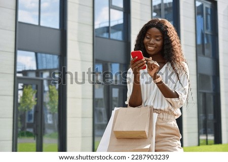 Happy young black African woman customer shopper standing on city street holding shopping bags using cell phone mobile app buying fashion sale clothes online in ecommerce digital store on smartphone.