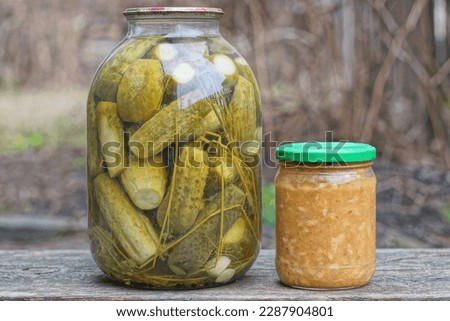  two glass jars with green pickled cucumbers and pickled salads stand on a gray wooden table in the street