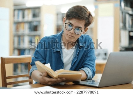 Young male college student wearing eyeglasses and in casual cloths studying and reading books using laptop at library for research or school project.