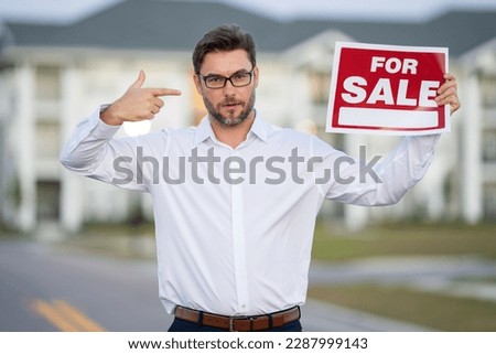 Realtor agent is a realtor with sign for sale in hand against the background on new apartment home background. Realtor in shirt, outdoor portrait. Realtor offering new home. Property concept.