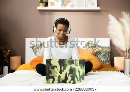 Photo of smiley delighted african american dark skinned woman wearing headphones sitting on bed in front of computer smiling with beautiful white teeth.