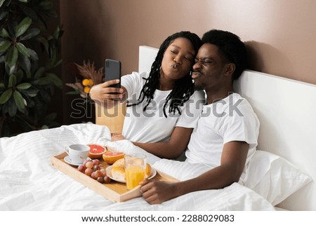 Happy black couple lying in bed having breakfast beautiful African American wife taking selfie pictures of two posing with different emotions.