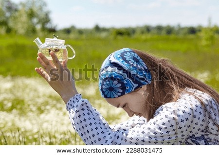 Woman holding transparent teapot with herbal tea. Collects fresh green herbs harvest and make healthy tea. Blurred image, selective focus