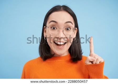 Eureka. Excited asian woman in prescribed glasses, raises finger up, has an idea and smiles, suggests smth, stands over blue background.