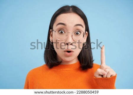 Portrait of smiling brunette asian woman in glasses, raises one finger, eureka sign, pitching an idea, has revelation, stands over blue background.
