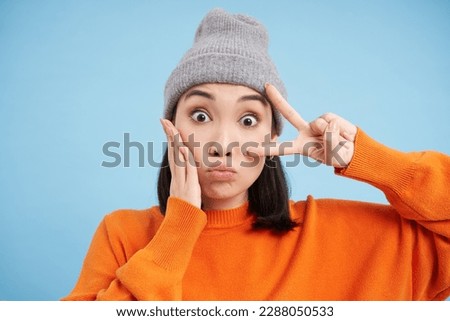 Close up portrait of cute girl in beanie, puckets lips and shows peace sign, posing kawaii against blue background.