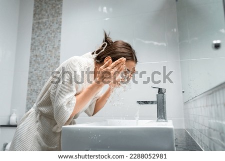 Asian beautiful woman washing her clean face with facial foam and water. Attractive female in bathrobe washing face for healthy beauty treatments and skin care then looking at the mirror in bathroom. Royalty-Free Stock Photo #2288052981