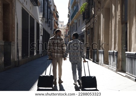 A young gay male couple walks down a street with their suitcases. The couple goes on a trip. The photo is taken from behind. Vacation and travel concept. Royalty-Free Stock Photo #2288053323