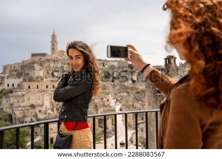 Young couple of woman friends taking pictures as souvenir with smartphone. Matera city in the background.