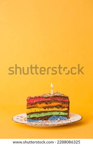 Rainbow birthday cake decorated with a festive candle and confetti. Confectionery. Multicolored dessert in the color of the rainbow on a yellow background.