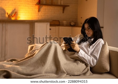 A young woman lies on a couch wrapped in a blanket and holds a digital tablet in her hands. A young brunette spends her evening watching funny videos on a digital tablet. Royalty-Free Stock Photo #2288094315