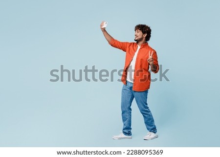 Full body fun young Indian man wear orange red shirt white t-shirt doing selfie shot on mobile cell phone post photo on social network show v-sign isolated on plain pastel light blue cyan background