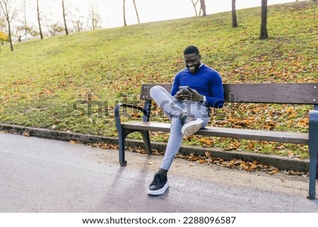 Young African man is sitting on a park bench wearing a casual blue sweater and glasses, absorbed in chatting on his mobile phone. The lighting is good, illuminating his features and the surroundings.