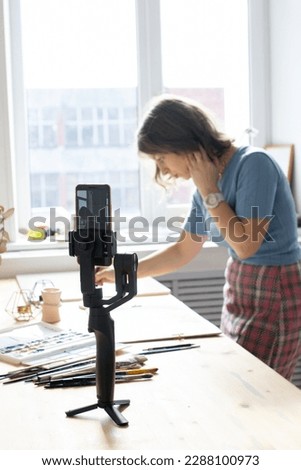 Young woman painting a picture with watercolor at wooden table, aquarelle process, girl sitting at the table and create picture, blogger filming video and making content