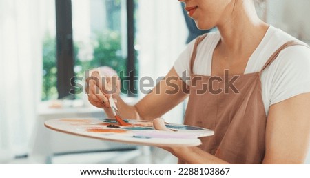 Close-up youth Asia lady wear apron holding palette color use brush painting on canvas create artwork in cozy workshop at home. Contemporary Painter Abstract Modern Art, Creativity and people concept.