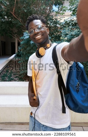 Vertical photo of a young student guy wearing headphones and carrying a file folder taking a selfie of him looking at camera smiling. Break-time outside campus patio, coming out of classroom.