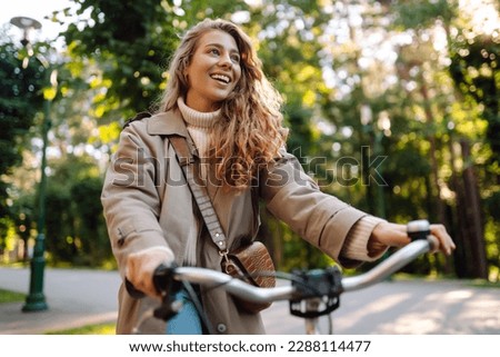 Smiling woman with curly hair in a coat rides a bicycle in a sunny park. Outdoor portrait. Beautiful woman enjoys nature. Lifestyle. Relax, nature concept.  Royalty-Free Stock Photo #2288114477