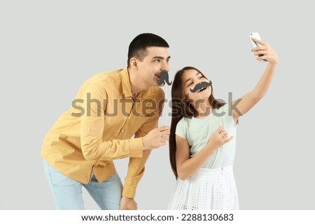 Portrait of father and his little daughter with paper mustache taking selfie on light background