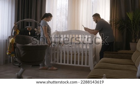 Happy parents stand near the child crib with little baby at cozy home. Father takes a picture or video of newborn daughter or son using smartphone. Concept of childhood, parenthood, love and family.