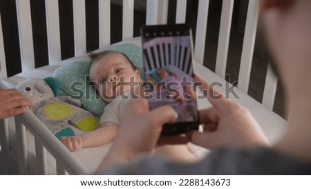 Father takes a picture or video of newborn playful daughter or son using smartphone. Parents stand near the child crib with little baby in bodysuit. Concept of childhood, parenthood, love and family.