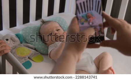 Parents stand near the child crib with little baby in bodysuit. Couple takes a picture or video of newborn adorable daughter or son on mobile phone. Concept of childhood, parenthood, love and family.