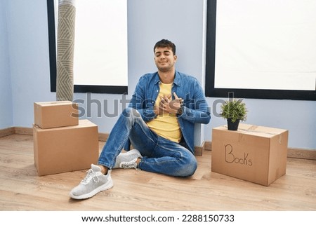 Young hispanic man sitting on the floor at new home smiling with hands on chest with closed eyes and grateful gesture on face. health concept. 