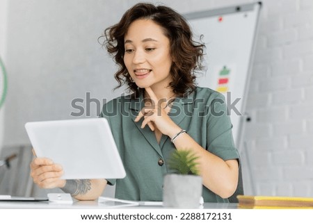 Tattooed speech therapist pointing at chin during video call on digital tablet in consulting room Royalty-Free Stock Photo #2288172131