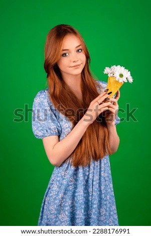 Beautiful attractive red-haired young girl in blue dress holding spring ice cream with daisy flowers posing on green background isolated.