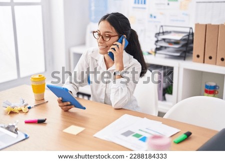 Young beautiful hispanic woman business worker using touchpad talking on smartphone at office