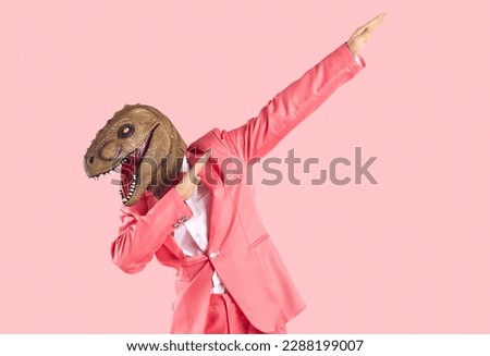 Cheerful young man in dinosaur rubber mask makes dab dance gesture isolated on pink background. Man with lizard mask on his head dressed in formal pink suit is having fun and shows dabbing movement. Royalty-Free Stock Photo #2288199007