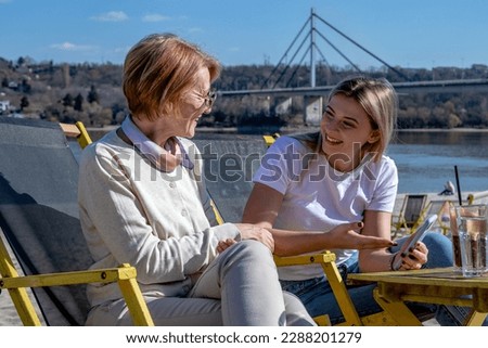 Mother and daughter spend quality time by the river together, bonding, and drinking coffee. The daughter shows her mum photos of her new family.