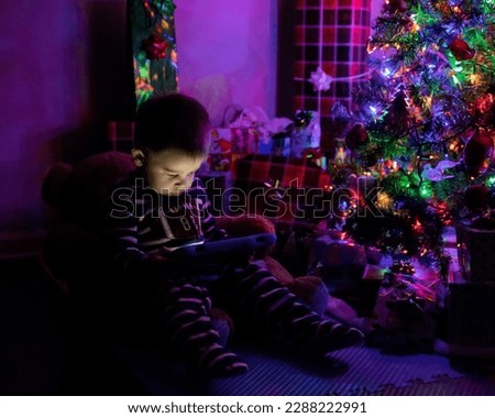 Young boy in pajamas sitting next to Christmas tree on Christmas eve playing on tablet waiting for presents to open